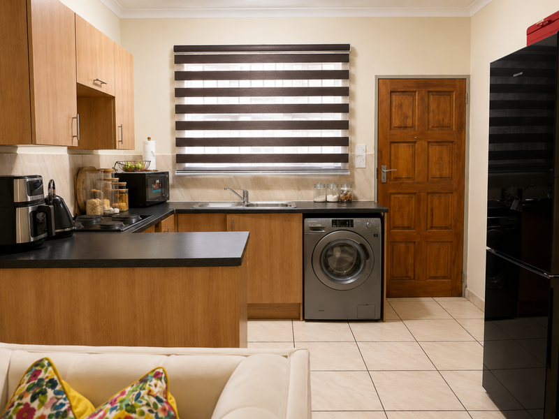A functional kitchen featuring light wood cabinetry, dark countertops, tiled flooring, and a built-in washing machine, complemented by a classic timber door.