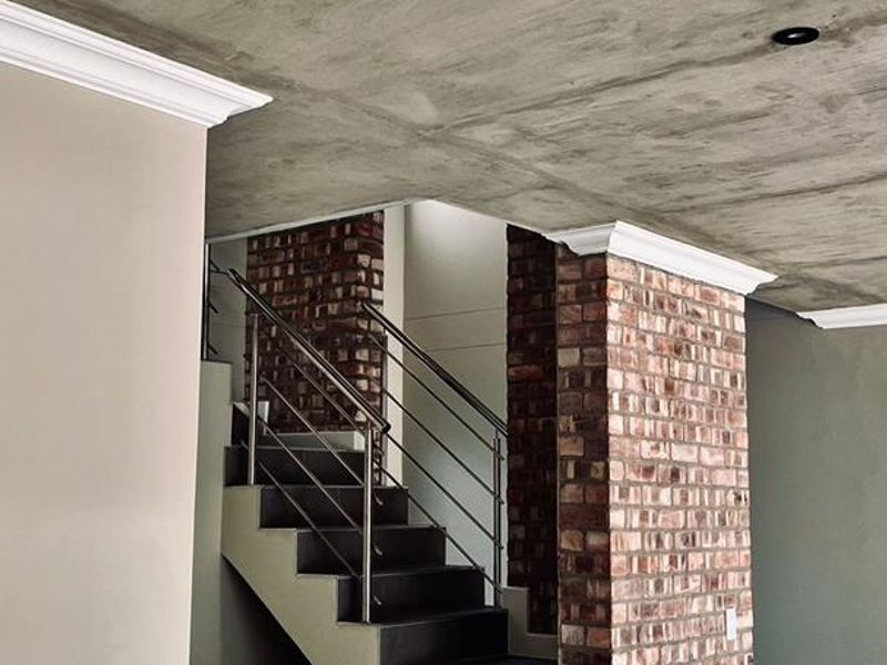 A modern hallway featuring an exposed concrete ceiling, striking brick columns, and a tiled staircase with a sleek stainless steel balustrade.