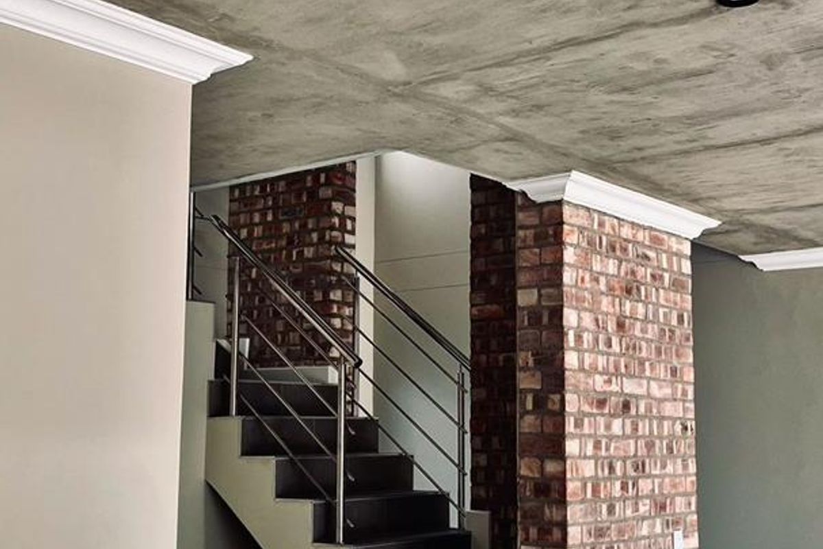 A modern hallway featuring an exposed concrete ceiling, striking brick columns, and a tiled staircase with a sleek stainless steel balustrade.