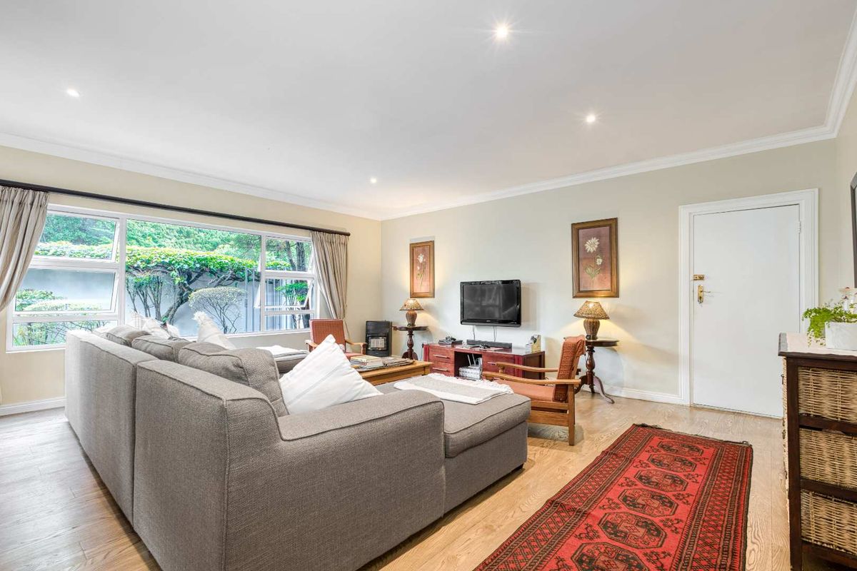 A bright and comfortable living room featuring large windows with garden views, recessed downlighting, and light-toned wood-look flooring.