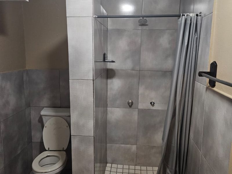 A functional bathroom featuring a walk-in shower with tiled surround and a standard toilet, finished with neutral grey wall and floor tiles.