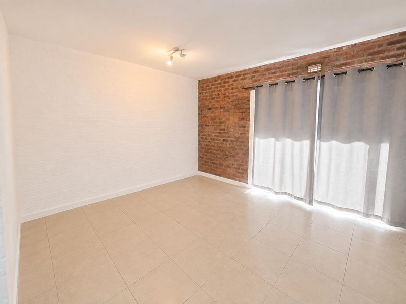 A bright, empty living room featuring a characterful exposed brick wall, neutral tiled flooring, and a large sliding door providing natural light.