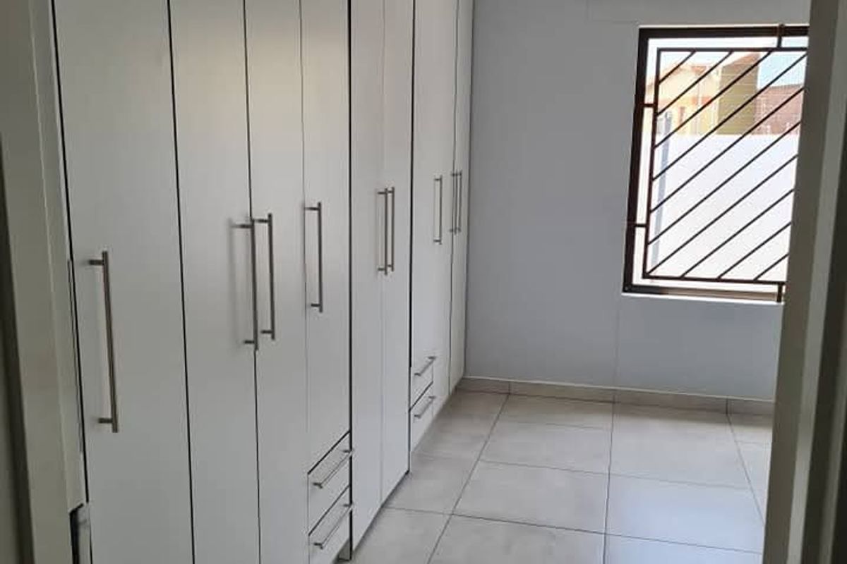 A bright bedroom featuring extensive floor-to-ceiling built-in cabinetry, neutral tiled flooring, and a window fitted with security bars.