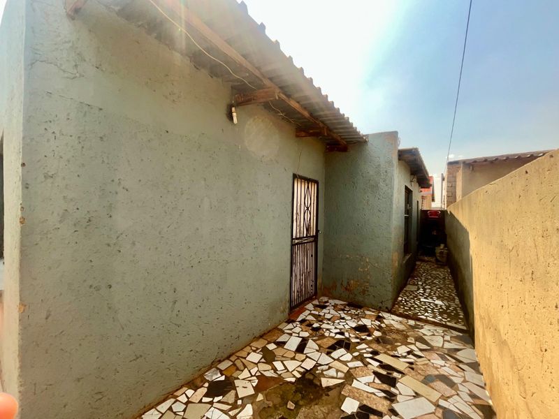 A narrow rear exterior passage featuring a security gate, corrugated roofing, and decorative crazy paving flooring with visible wear to the wall finishes.