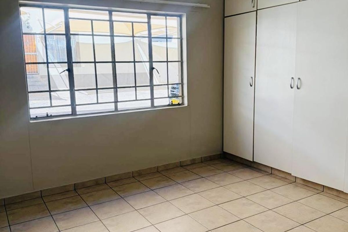 A clean, empty bedroom featuring neutral tiled flooring, built-in storage cupboards, and a window fitted with security bars.