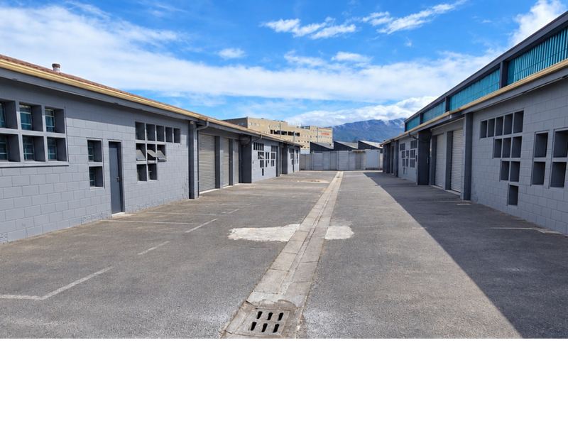 Exterior view of an industrial complex featuring concrete block buildings, asphalt driveway, and multiple roller shutter access points.