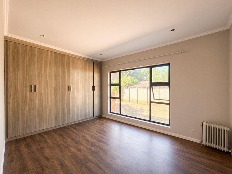 A bright, empty bedroom featuring modern wood-grain built-in cupboards, laminate flooring, a large window, and a wall-mounted radiator.