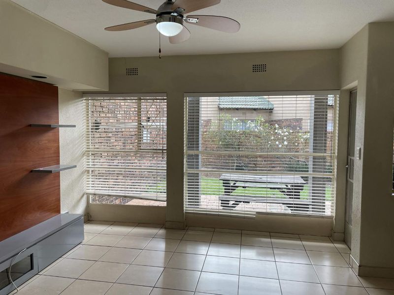 A living room featuring tiled flooring, a built-in media unit, a ceiling fan, and large windows overlooking a private garden area.
