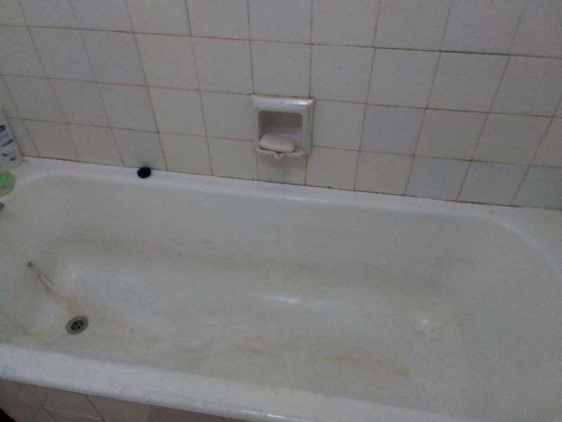 A dated bathroom featuring a built-in bathtub with integrated soap dish and ceramic wall tiling, showing significant signs of wear and surface staining.