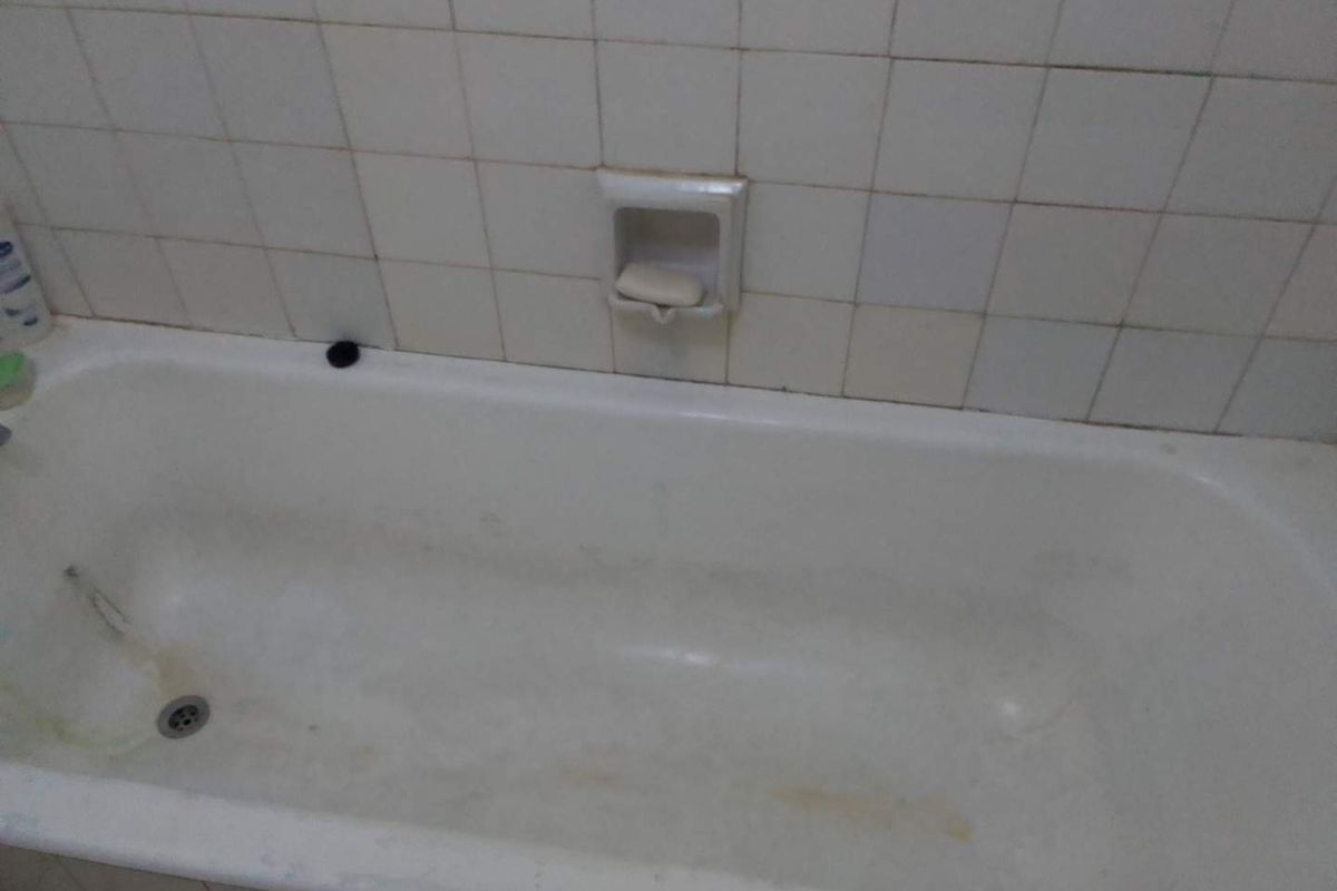A dated bathroom featuring a built-in bathtub with integrated soap dish and ceramic wall tiling, showing significant signs of wear and surface staining.