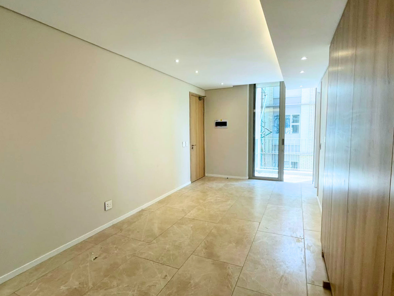 A bright, modern, and empty living area featuring neutral stone tile flooring, recessed ceiling lighting, and integrated cabinetry with access to a balcony.