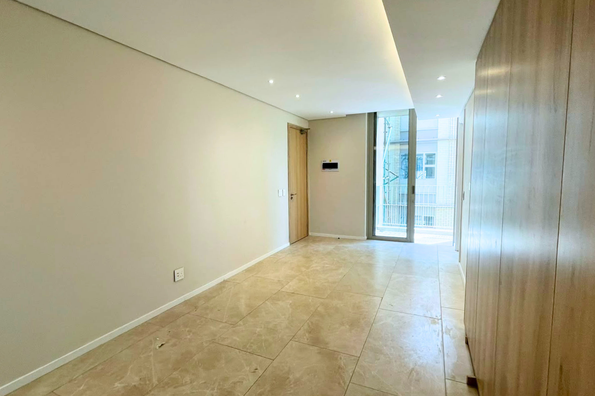 A bright, modern, and empty living area featuring neutral stone tile flooring, recessed ceiling lighting, and integrated cabinetry with access to a balcony.