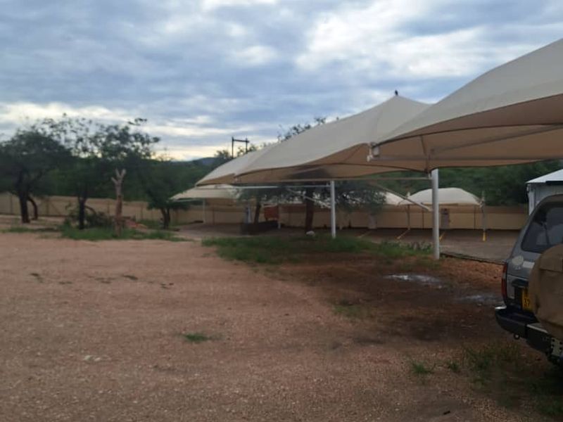 An outdoor industrial parking area featuring multiple shade sail structures over a gravel surface, enclosed by perimeter fencing.