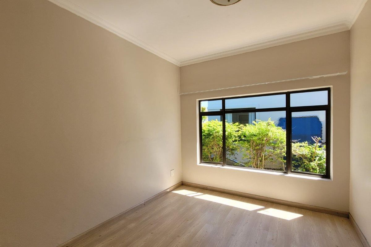 A bright, empty bedroom featuring light-toned laminate flooring, neutral wall finishes, and a large window with dark frames providing natural light.