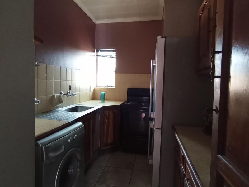 A compact kitchen featuring dated wooden cabinetry, tiled backsplash, stainless steel sink, and space for a freestanding stove and laundry appliance.