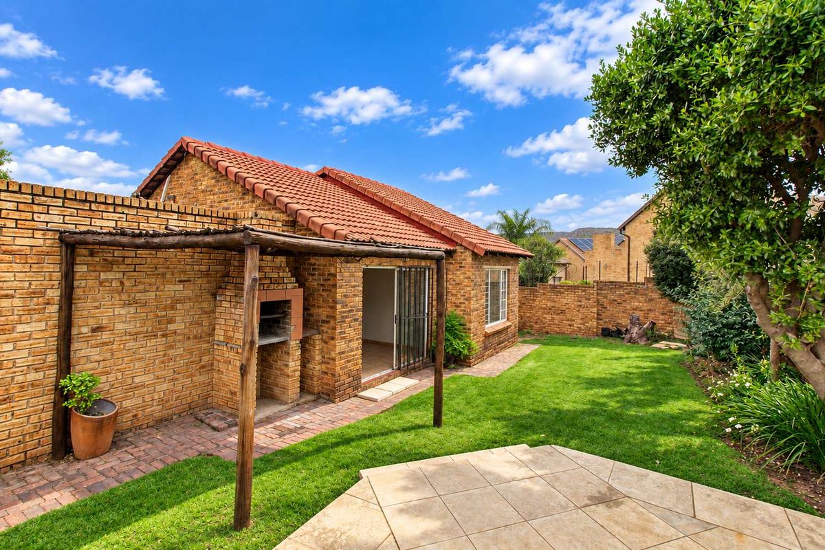 Rear exterior view of a townhouse featuring a brick facade, tiled roof, paved patio area, built-in braai, and a well-maintained garden.