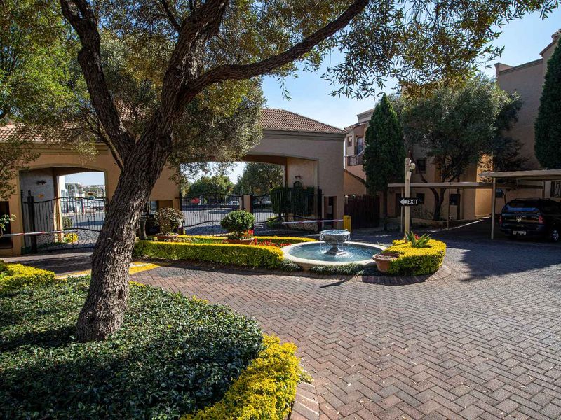 A well-maintained gated entrance to the apartment complex featuring paved driveways, landscaped garden beds, and a central decorative fountain.
