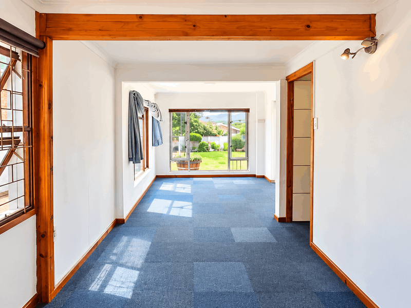An empty, sunlit living area featuring exposed timber beams, blue carpet tiles, and large windows providing views of the rear garden.