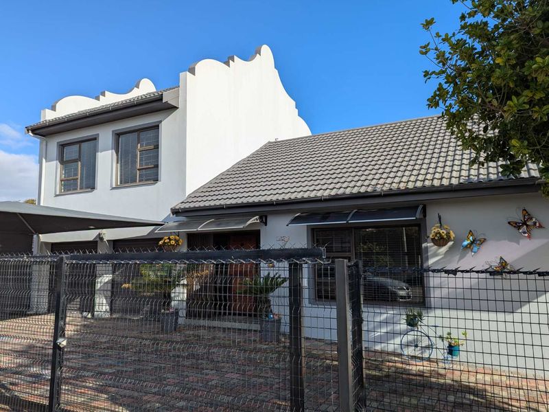 Front elevation of a two-story house featuring a gabled parapet, tiled roof, paved driveway, and secure wire mesh perimeter fencing.