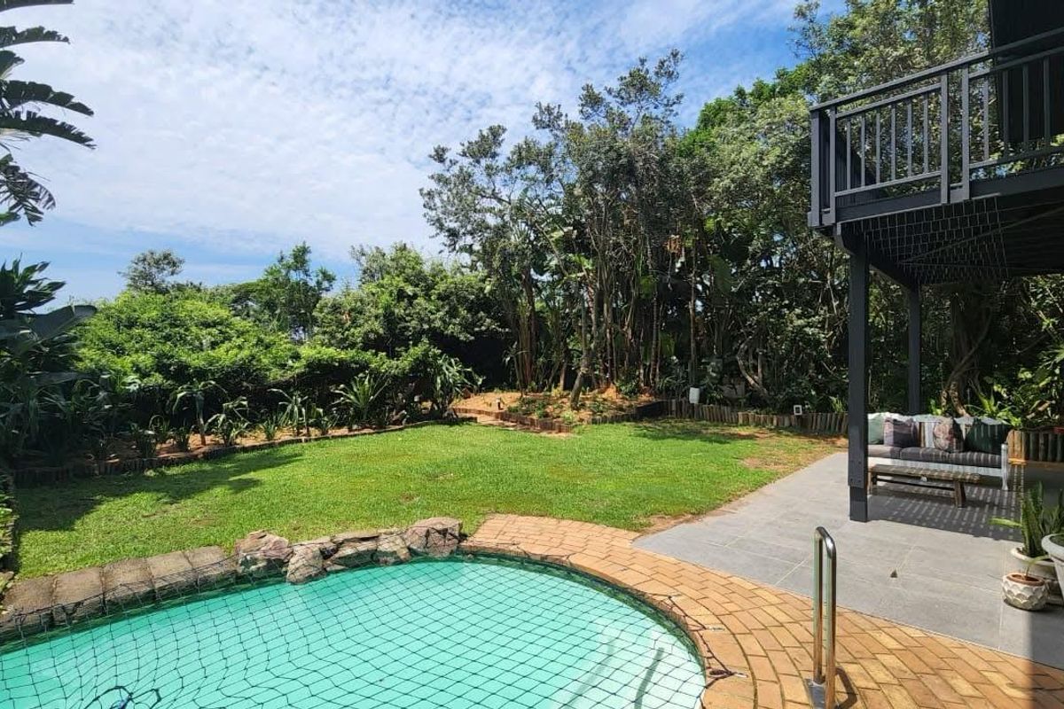 A private rear garden featuring a swimming pool with safety net, paved patio area, and a raised balcony deck overlooking the lush green space.