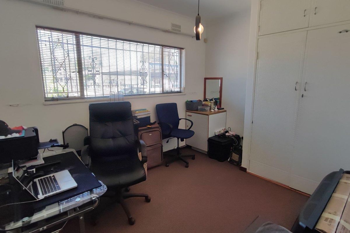 A functional bedroom or home office space featuring built-in cupboards, carpeted flooring, and a large window with security bars.
