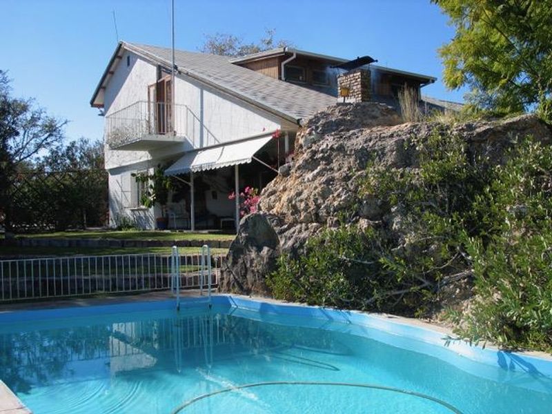 Exterior view of a residential property featuring a swimming pool, a natural rock formation, and a multi-level house with a balcony and chimney.