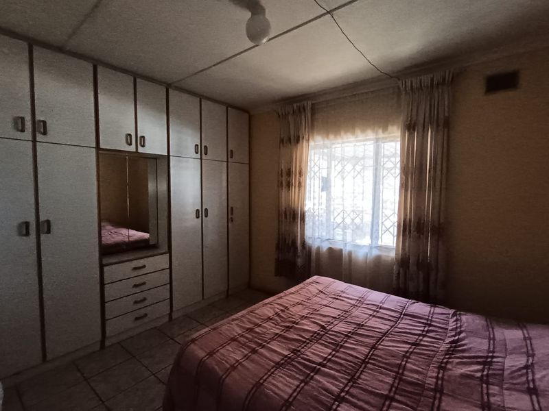 A bedroom featuring built-in cupboards, tiled flooring, and security bars on the window, requiring cosmetic updates to modernize the space.