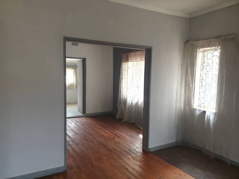 An empty living area featuring original hardwood flooring, large windows with security bars, and an open archway leading to an adjacent room.