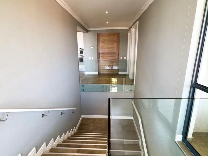 A bright, modern hallway featuring a tiled staircase with a sleek glass balustrade, crown molding, and a solid timber entrance door.