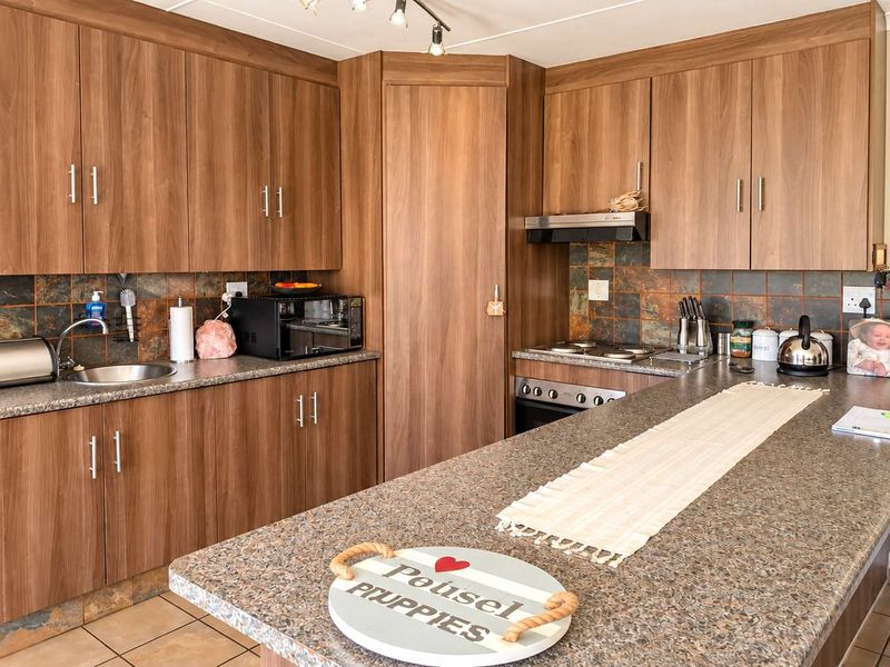 Functional kitchen featuring wood-look cabinetry, stone-effect countertops, built-in oven, electric hob, and tiled backsplash with neutral floor tiles.