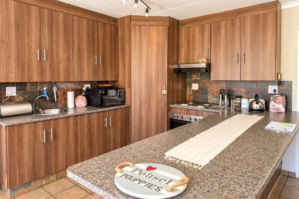 Functional kitchen featuring wood-look cabinetry, stone-effect countertops, built-in oven, electric hob, and tiled backsplash with neutral floor tiles.