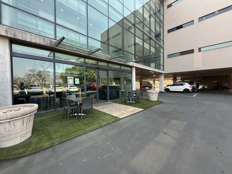 Modern office building entrance featuring a glass facade, glass canopy, and a small outdoor seating area with artificial turf.