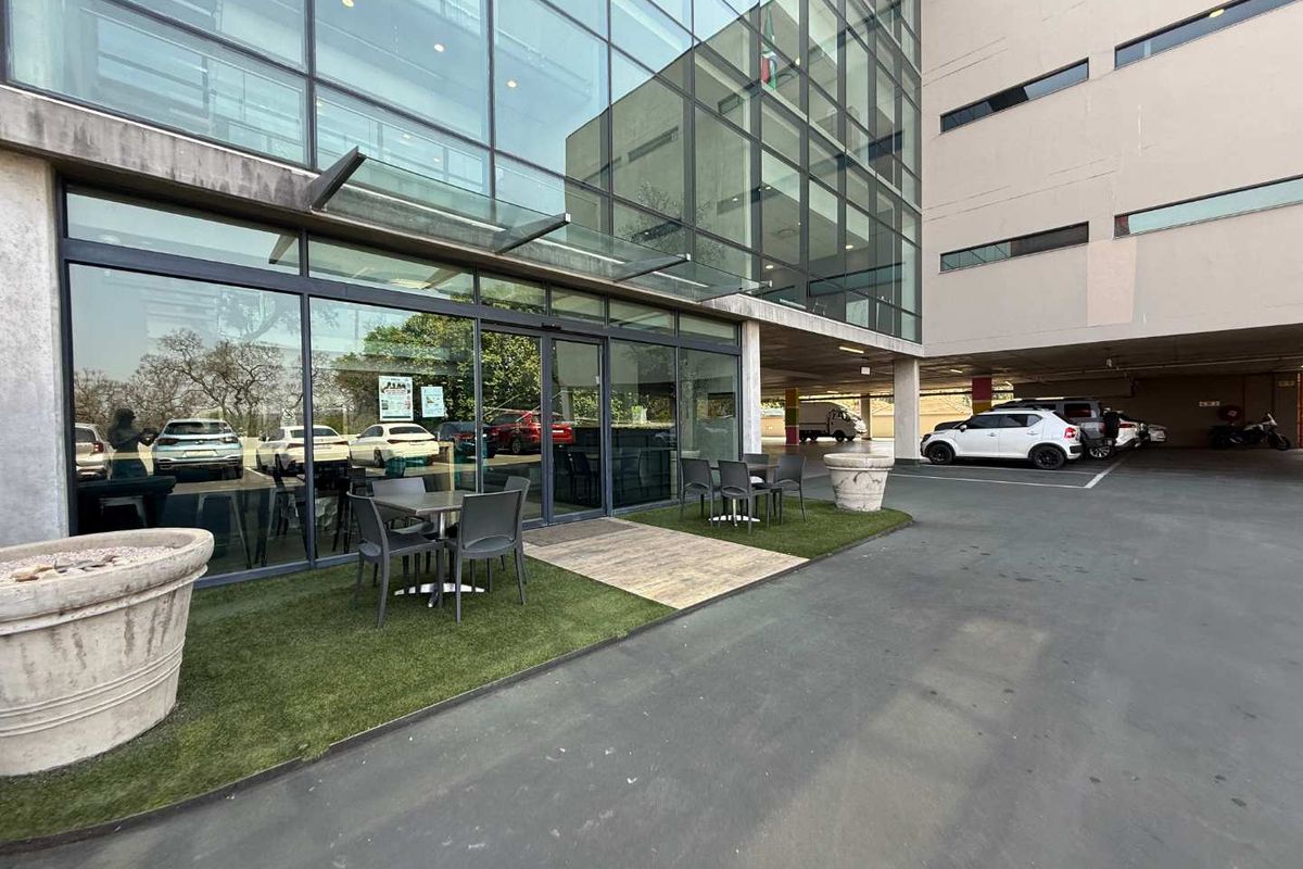 Modern office building entrance featuring a glass facade, glass canopy, and a small outdoor seating area with artificial turf.