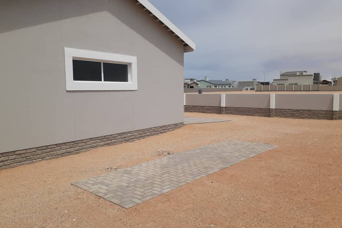 A view of the rear exterior of the house featuring a paved walkway, perimeter boundary wall, and low-maintenance sandy ground cover.