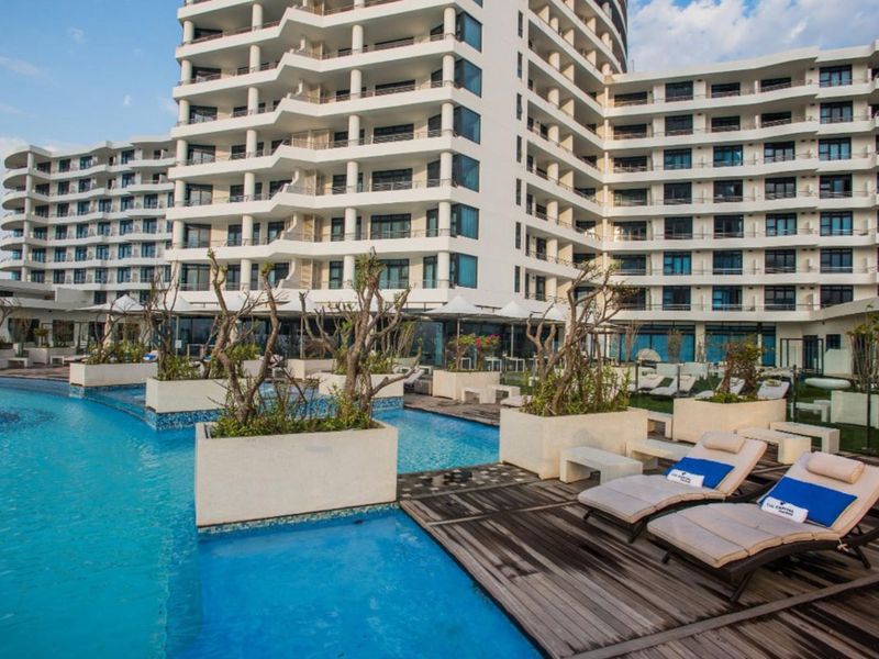 A well-maintained outdoor swimming pool area featuring timber decking, built-in planters, and a view of the modern multi-story apartment building facade.