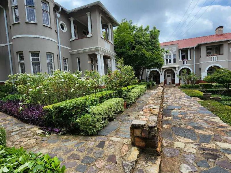 A well-maintained exterior rear view featuring a stone-paved walkway, landscaped garden beds, and classic architectural details including columns and a balcony.