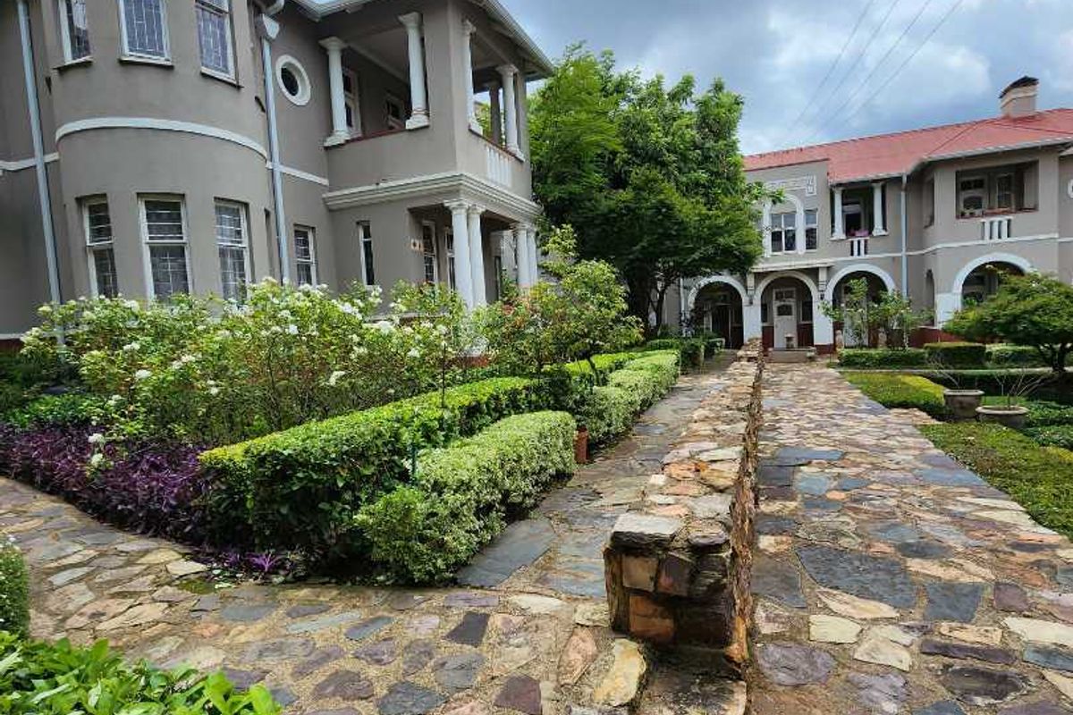 A well-maintained exterior rear view featuring a stone-paved walkway, landscaped garden beds, and classic architectural details including columns and a balcony.