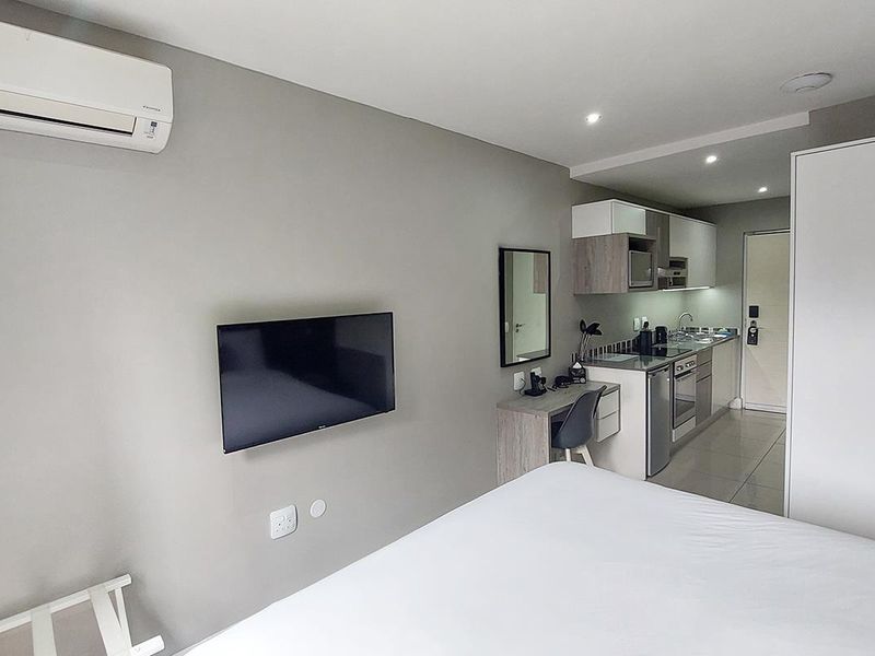Modern open-plan studio apartment featuring a compact kitchenette with built-in appliances, tiled flooring, and a wall-mounted air conditioning unit.