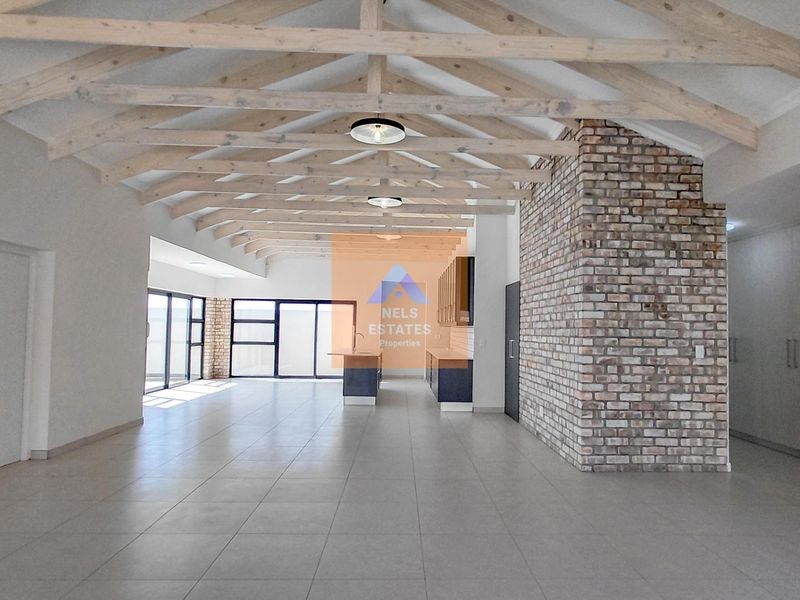 Spacious open-plan living area featuring exposed timber roof trusses, a striking brick feature wall, tiled floors, and modern kitchen cabinetry with an island.