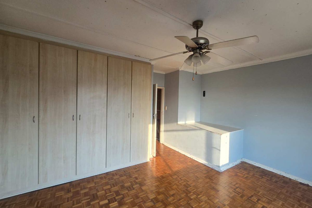 A bedroom featuring parquet flooring, built-in wooden cupboards, and a ceiling fan, offering a functional space with potential for cosmetic updates.