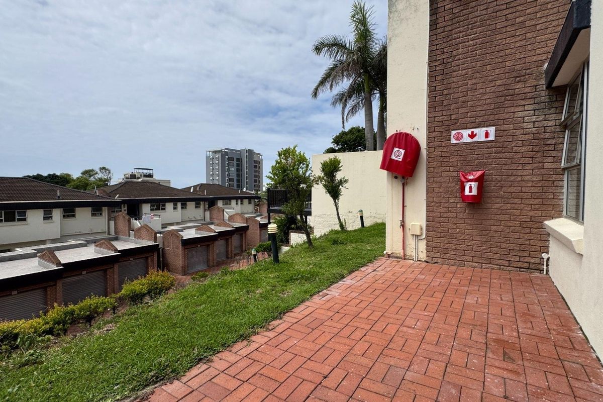 An elevated rear exterior area featuring brick paving, a grass verge, and installed fire safety equipment against the building facade.