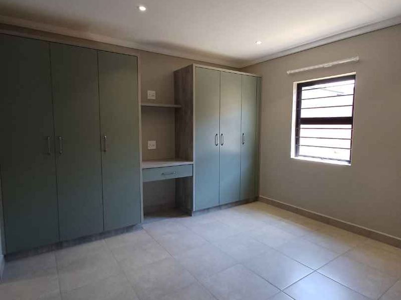 A clean, modern bedroom featuring neutral tiled flooring, recessed lighting, and extensive built-in cabinetry with an integrated desk area.