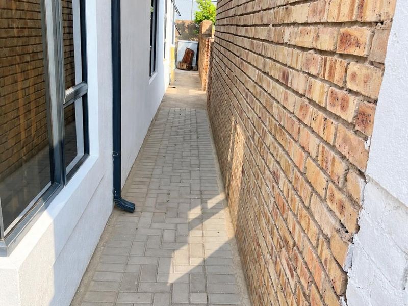 A narrow paved side passage between the house exterior and a brick boundary wall, featuring aluminium window frames and a drainage downpipe.