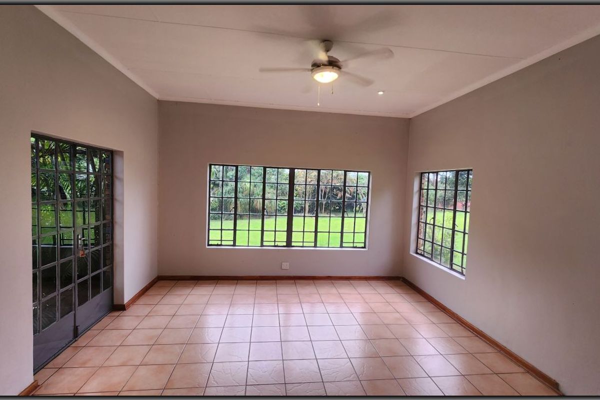 A spacious, empty living area featuring neutral walls, tiled flooring, a ceiling fan, and large steel-framed windows providing natural light and garden views.