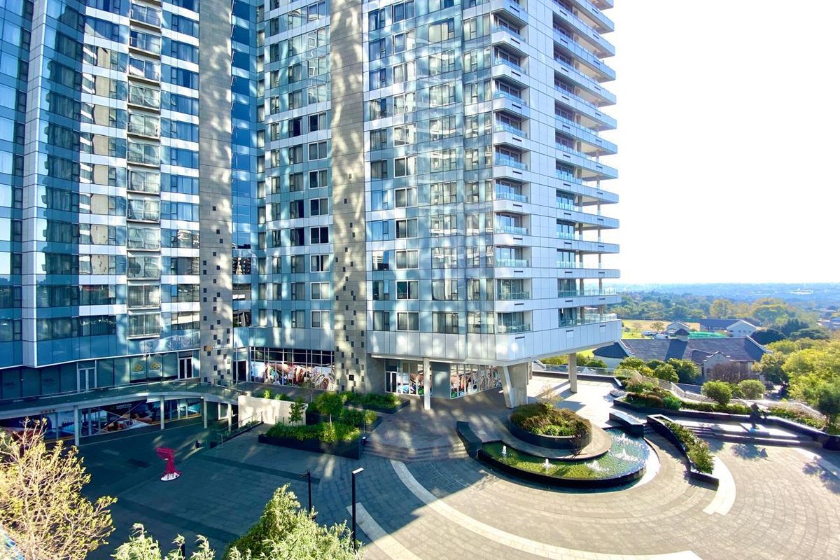 An elevated view of a modern high-rise apartment building featuring glass facades, balconies, and a landscaped courtyard with a central water feature.
