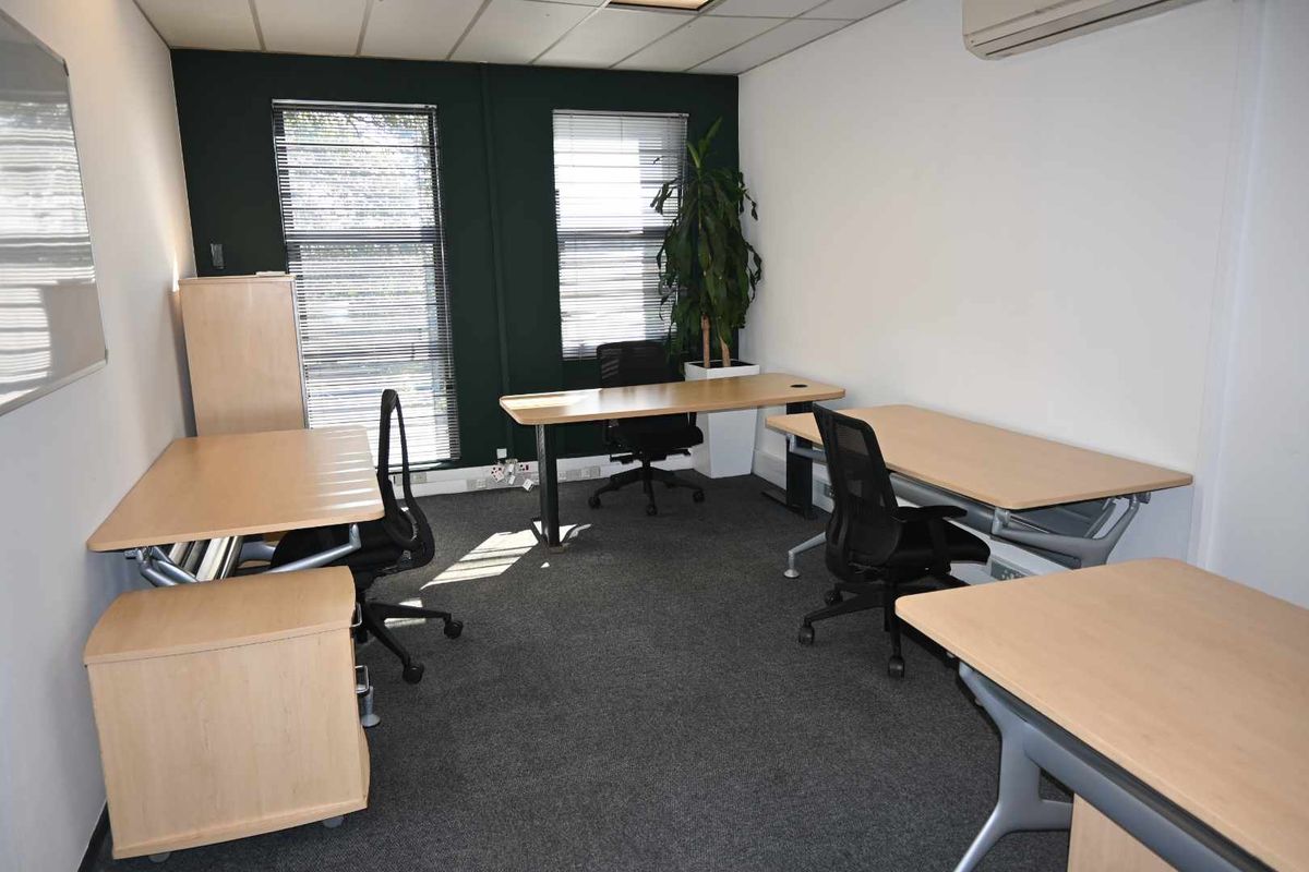 A functional office space featuring carpeted flooring, suspended ceiling, air conditioning, and large windows with venetian blinds.