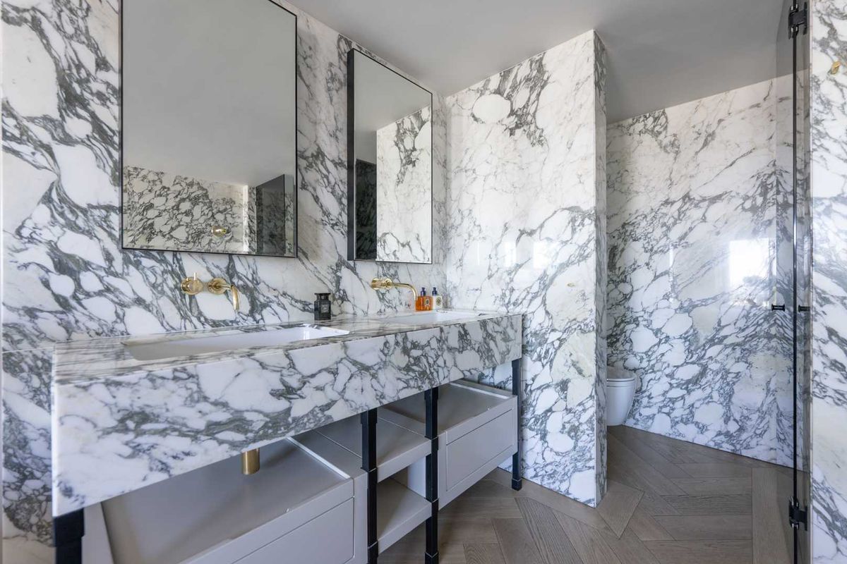 Luxurious modern bathroom featuring dual marble vanity sinks, wall-mounted gold fixtures, and elegant herringbone timber flooring.