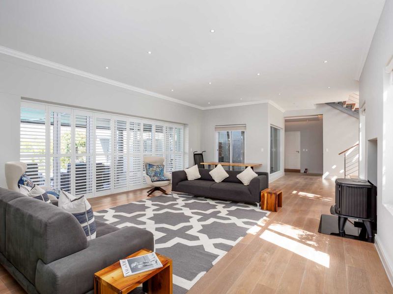 Spacious living room featuring light timber flooring, recessed downlights, plantation shutters, and a modern freestanding wood-burning fireplace, leading to an open staircase.