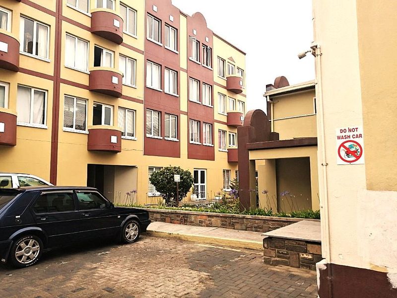 Exterior view of an apartment complex featuring a paved driveway, stone retaining wall, and multiple balconies on the building facade.