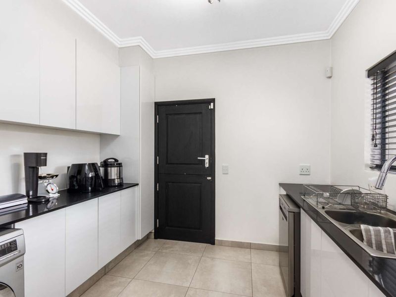 A modern, well-maintained scullery featuring sleek white cabinetry, dark stone countertops, a double basin sink, and tiled flooring.